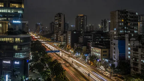 A Mumbai arterial road at night from above, traffic drawn out into light trails between lit office towers.