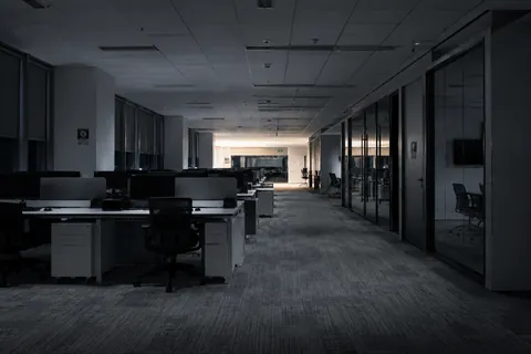 An unlit open-plan office at night, empty desks in rows and glazed meeting rooms along one side.