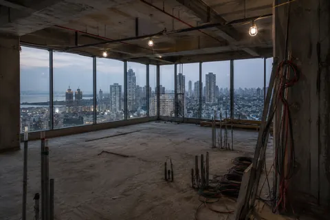 A bare concrete floor plate in an unfinished high-rise, services dropped from the slab and the city through open glazing.